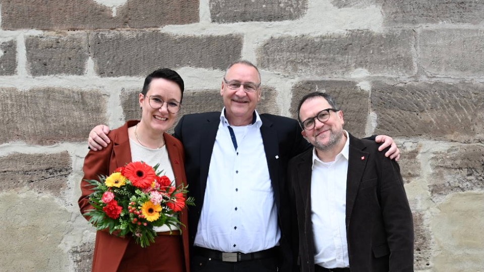 Die beiden neu gewählten Bevollmächtigten Jan Körper und Eva Wohlfahrt stehen vor einer Wand, zwischen ihnen steht der scheidende erste Bevollmächtigte Klaus-Dieter Winnerlein. Jan Körper hält einen Blumenstraß.