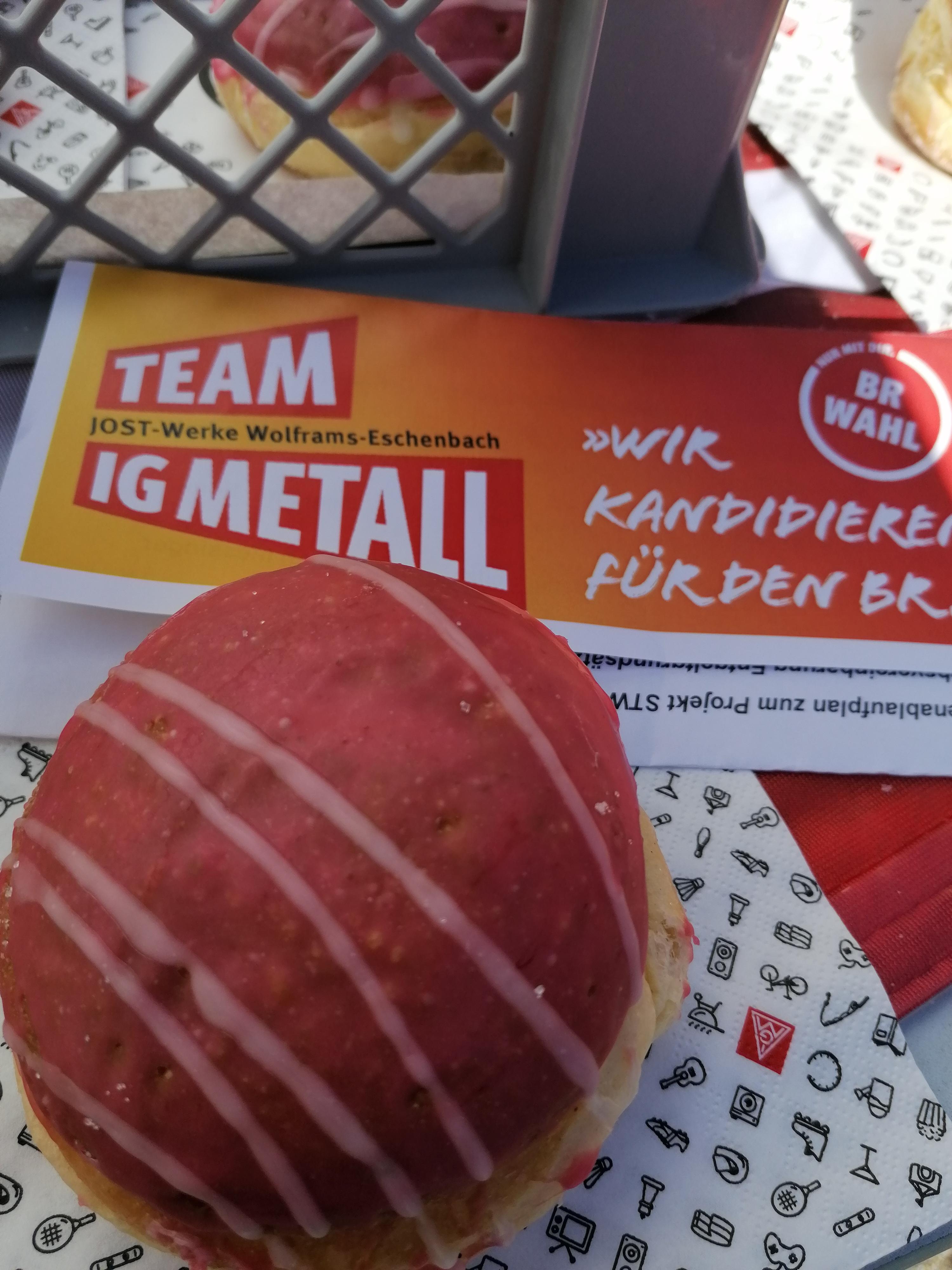 Ein rot-weißer Krapfen liegt gemeinsam mit einem Wahlflyer für die Kandidat*innen der IG Metall-Liste zur BR-Wahl 2022 und einer IG Metall-Serviette auf einem Tisch.