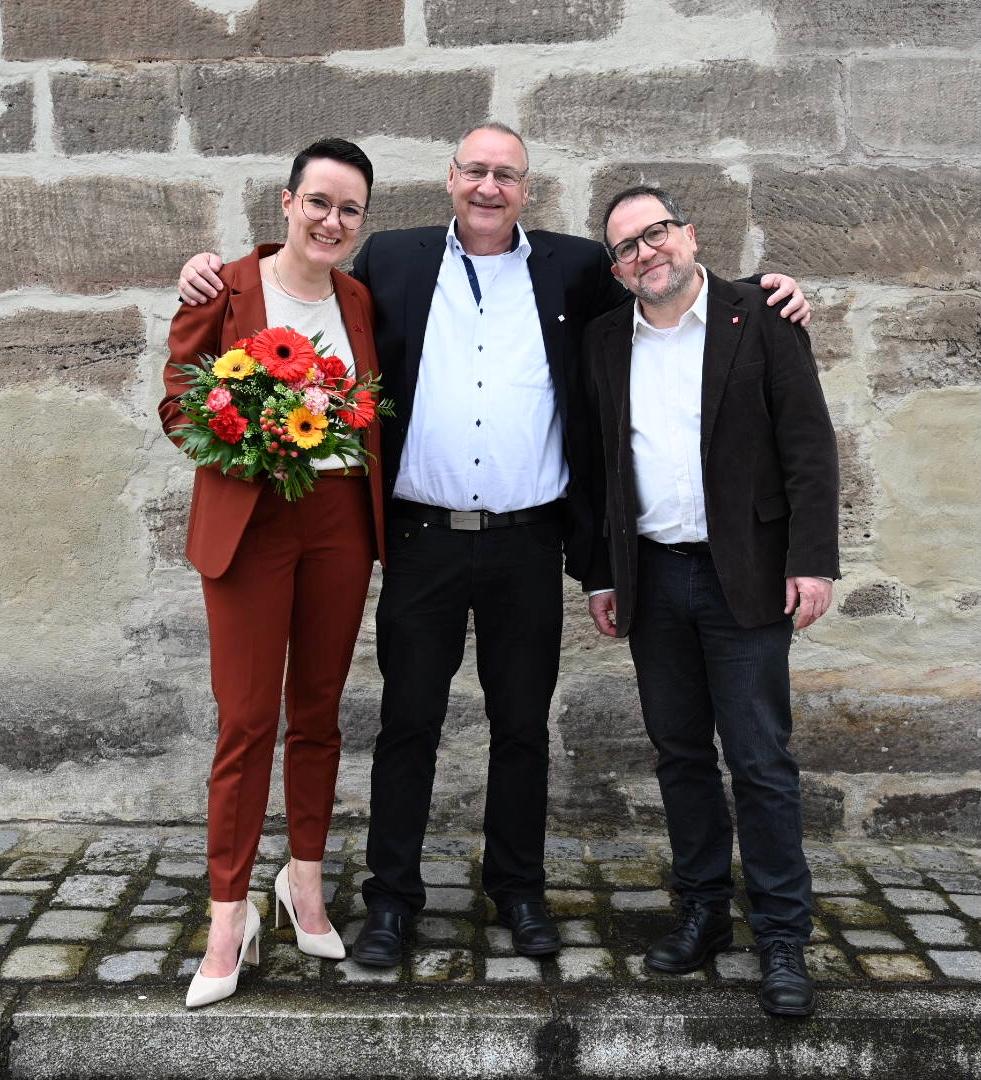 Die beiden neu gewählten Bevollmächtigten Jan Körper und Eva Wohlfahrt stehen vor einer Wand, zwischen ihnen steht der scheidende erste Bevollmächtigte Klaus-Dieter Winnerlein. Eva Wohlfahrt hält einen Blumenstraß.