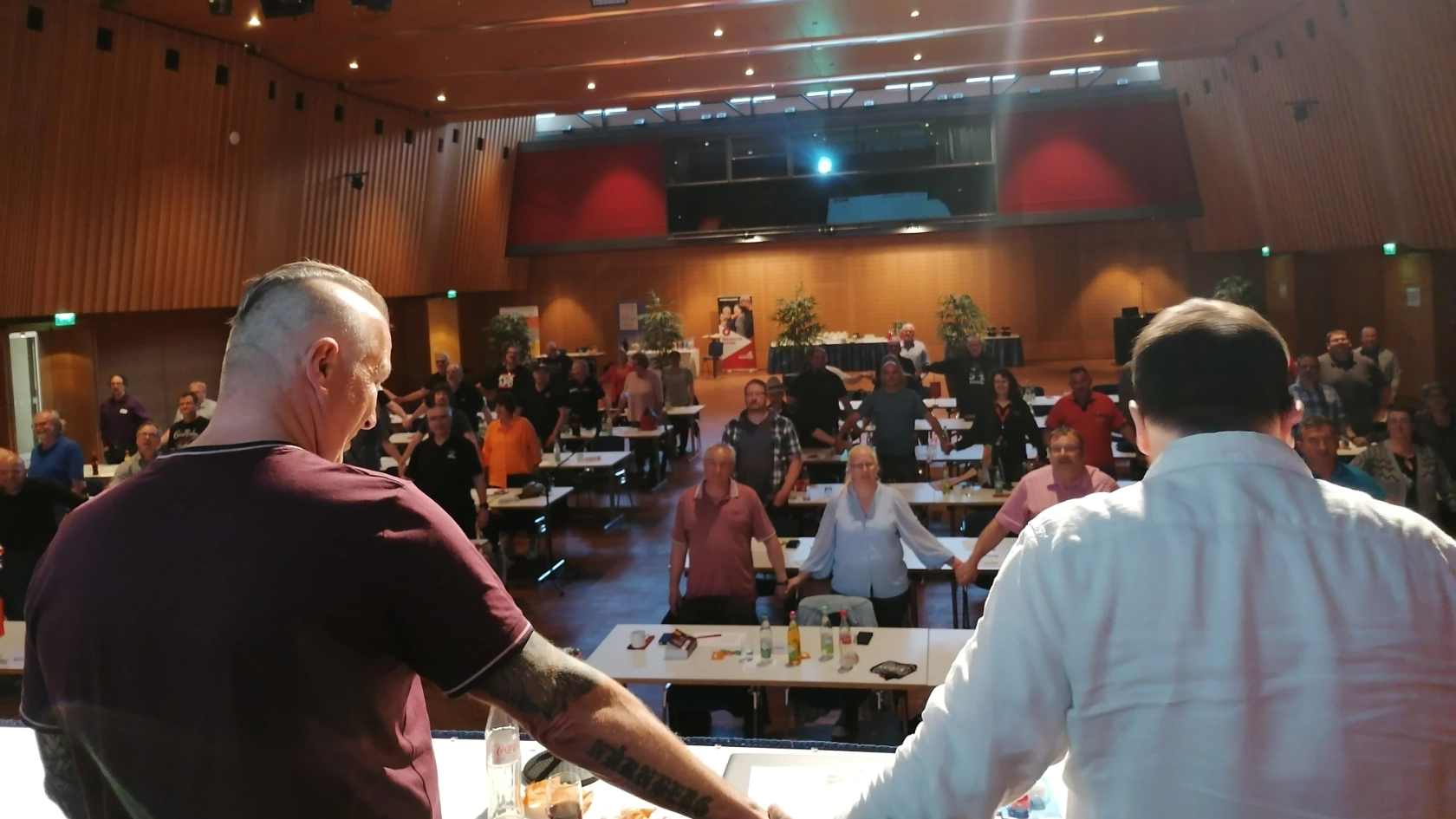 Zwei Kollegen halten sich an der Hand und sehen ins Plenum, das sich ebenfalls an der Hand hält.