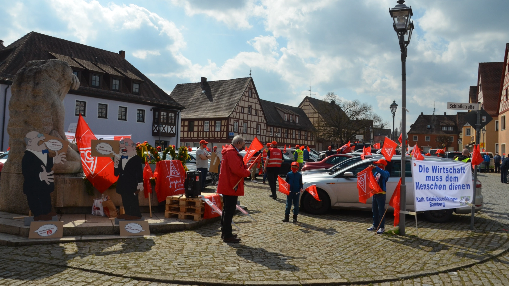 Menschen stehen mit IG Metall-Fahnen, Plakaten und geschmückten Autos bei einer Kundgebung auf einem Platz.