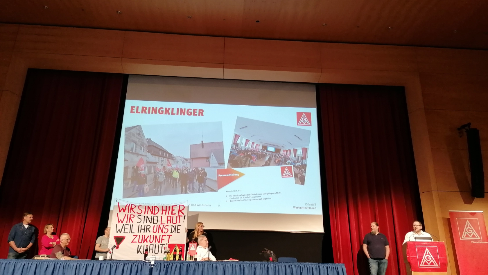 Die Kolleg*innen von Elring Klinger stehen mit einem Transparent auf der Bühne, im Hintergrund Bilder der letzten Demo gegen die Betriebsschließung. Jan Körper hält eine Rede.