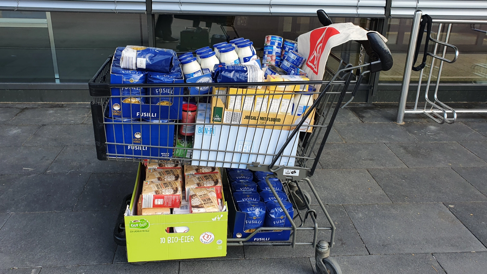 Ein voll gepackter Einkaufswagen steht vor einem Laden. In ihm befinden sich volle Kartonagen mit Mehl, Milch, Nudeln, Reis, Marmelade, Konserven, Salz und Öl als Spende für die Tafel Ansbach. Der Einkaufswagen ist mit einer IG Metall-Tasche geschmückt. 