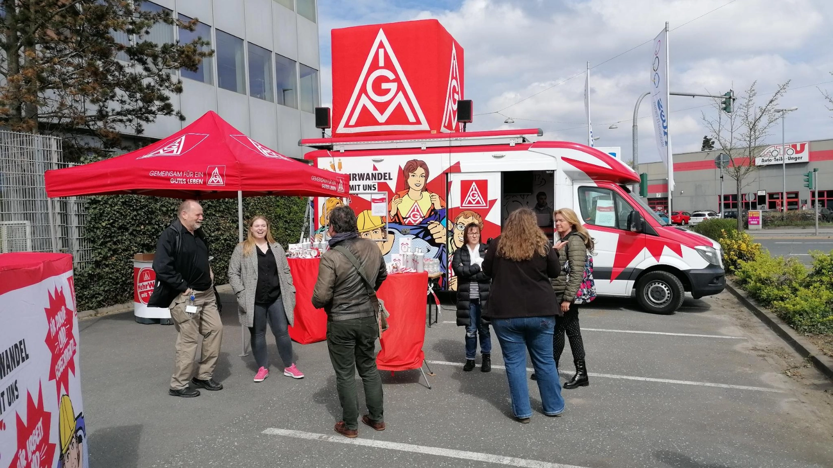Im Hintergrund ist der Roadshowtruck der IG Metall auf dem Werksgeländer der Firma Atos in Fürth zu sehen. Es weht die Fahne des Betriebes, Im Vordergrund sind Kolleg*innen zu sehen, die sich gemeinsam unterhalten und lachen. 