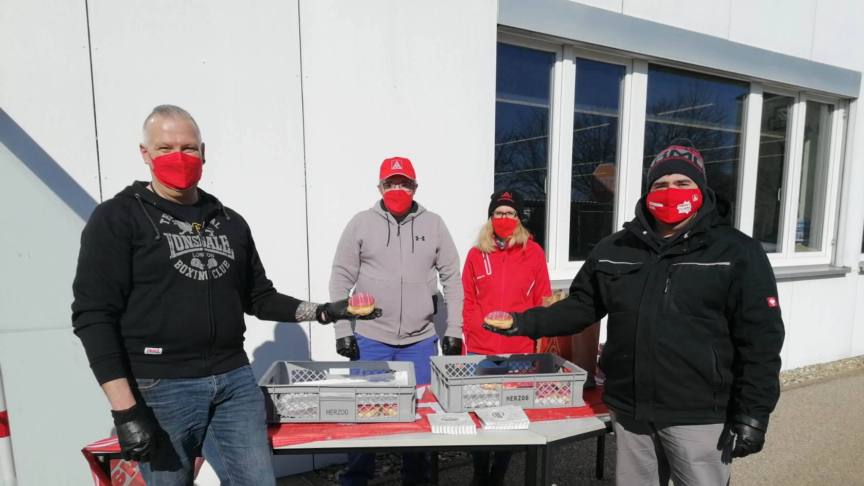 Vier Kolleg*innen der IG Metall-Liste stehen an einem Stand mit rot-weißen Faschingskrapfen und verteilen diese im Zuge der Betriebsratswahl bei Jost in Wolframs-Eschenbach