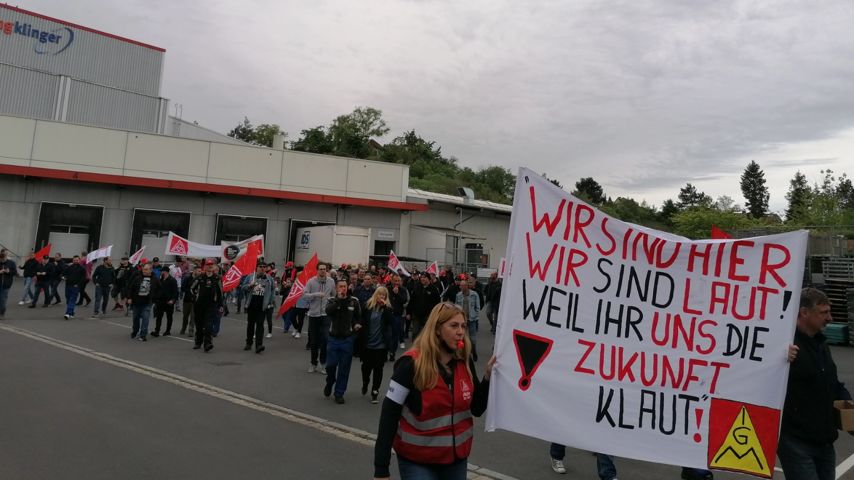 Die Beschäftigten von ElringKlinger laufen gemeinsam mit einem Demozug gegen die geplante Werksschließung durch Langenzenn. Sie halten ein Transparent mit der Aufschrift "Wir sind hier, wir sind laut, weil ihr uns die Zukunft klaut" hoch und tragen IG Metall-Westen.
