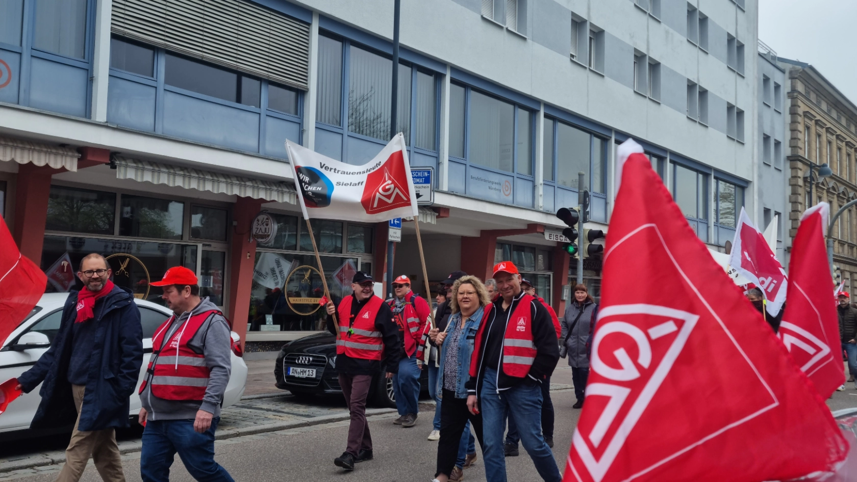Demozug läuft mit Transparenten und Bannern an einem mit IG Metall Fahnen geschmückten Auto vorbei.