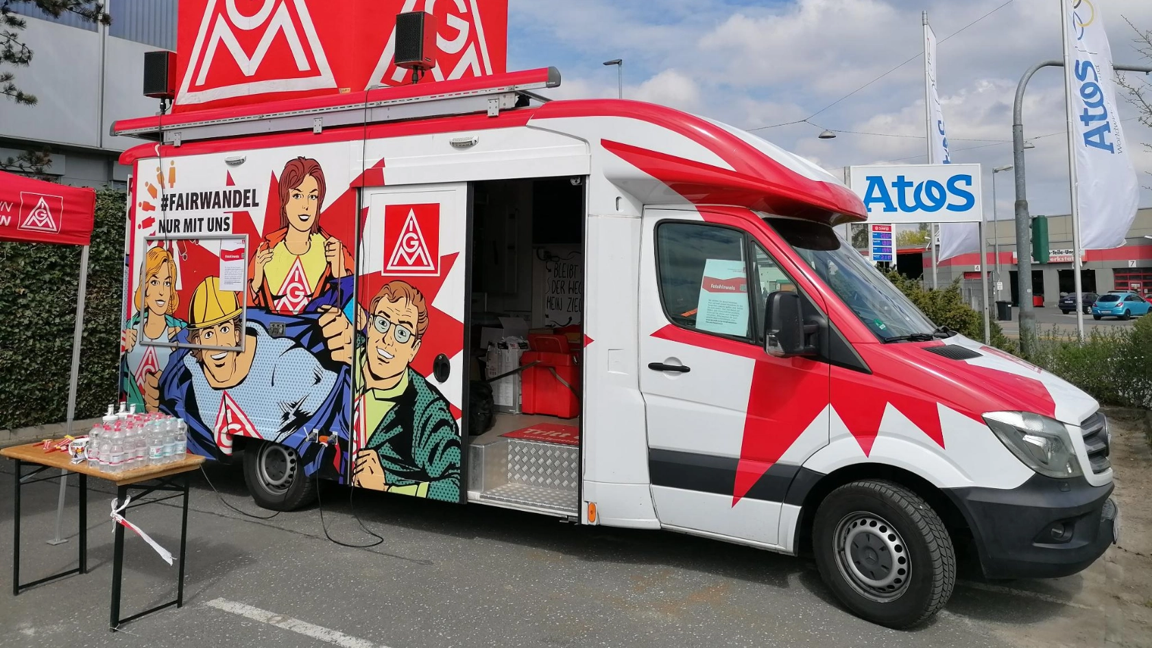 Ein rot-weißer IG Metall Truck steht auf dem Betriebsgeländer der Firma Atos in Fürth. Im Hintergrund sieht man die Firmenflagge wehen. Auf dem Truck steht ein großer IG Metall-Würfel.