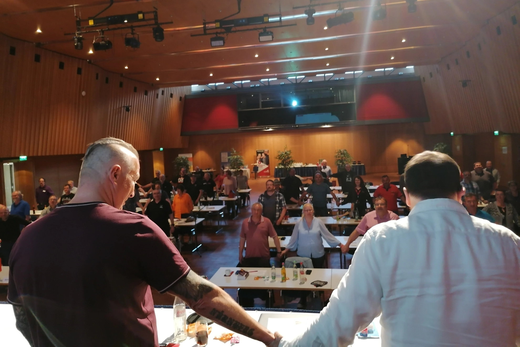 Zwei Kollegen halten sich an der Hand und sehen ins Plenum, das sich ebenfalls an der Hand hält.