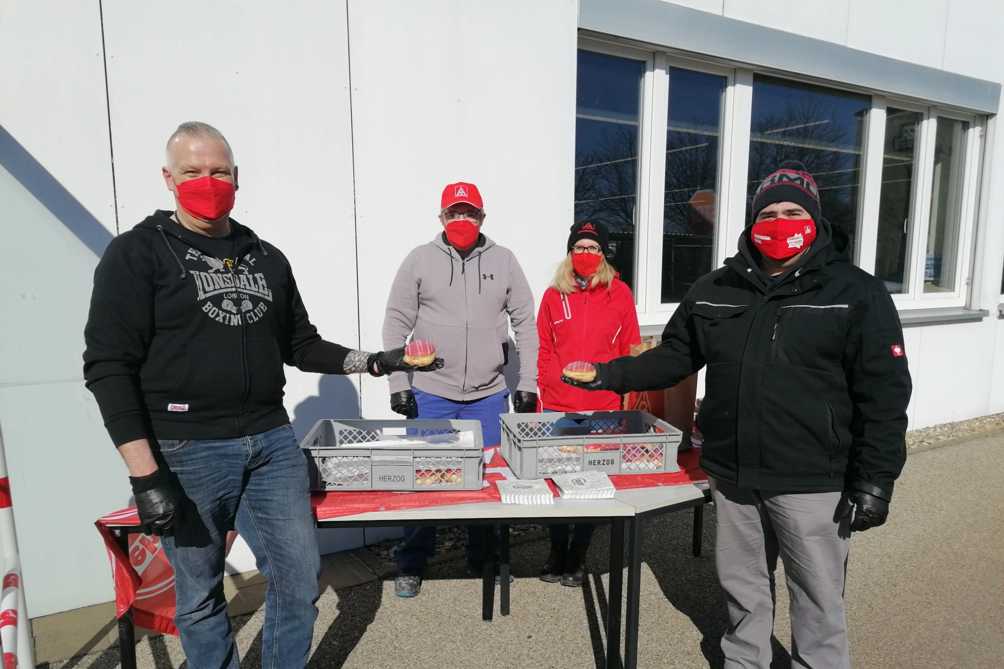Vier Kolleg*innen der IG Metall-Liste stehen an einem Stand mit rot-weißen Faschingskrapfen und verteilen diese im Zuge der Betriebsratswahl bei Jost in Wolframs-Eschenbach