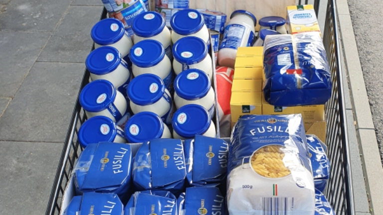 Ein Einkaufswagen steht vor einem Laden und ist gefüllt mit Grundnahrungsmitteln wie Nudeln, Reis, Mehl, Öl und Konserven und geschmückt mit einer IG Metall-Tasche. 