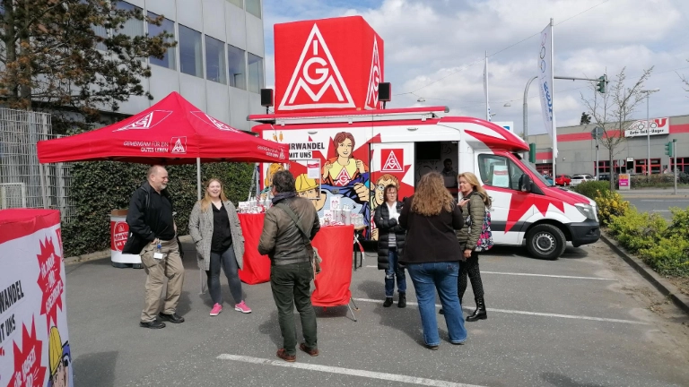 Im Hintergrund ist der Roadshowtruck der IG Metall auf dem Werksgeländer der Firma Atos in Fürth zu sehen. Es weht die Fahne des Betriebes, Im Vordergrund sind Kolleg*innen zu sehen, die sich gemeinsam unterhalten und lachen. 