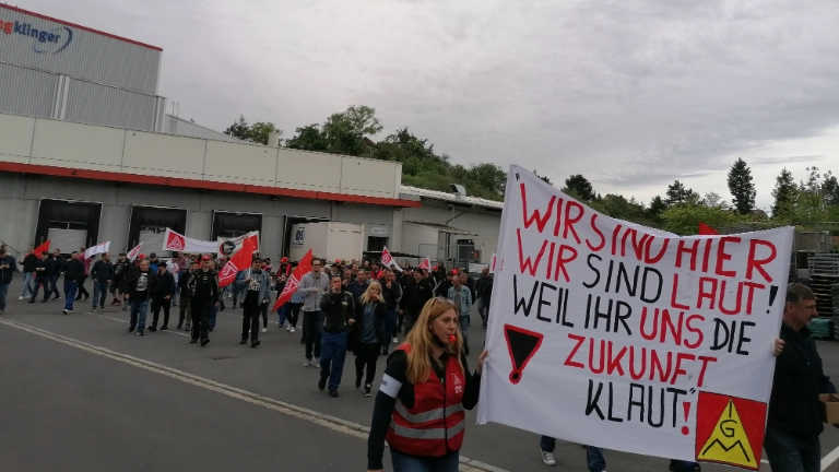 Die Beschäftigten von ElringKlinger laufen gemeinsam mit einem Demozug gegen die geplante Werksschließung durch Langenzenn. Sie halten ein Transparent mit der Aufschrift "Wir sind hier, wir sind laut, weil ihr uns die Zukunft klaut" hoch und tragen IG Metall-Westen.