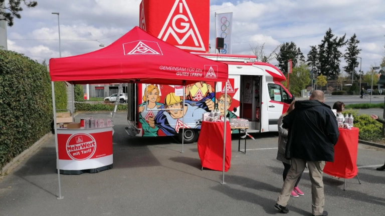 im Vordergrund ist ein roter IG Metall Pavillon zu sehen, im Hintergrund der rot-weiße Truck der Roadshow und die Flagge der Firma Atos. Es stehen Kolleg*innen vor dem Truck und unterhalten sich.